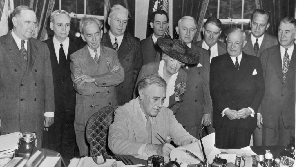 President Franklin D. Roosevelt seated at his desk signing the G.I. Bill as lawmakers stand around him